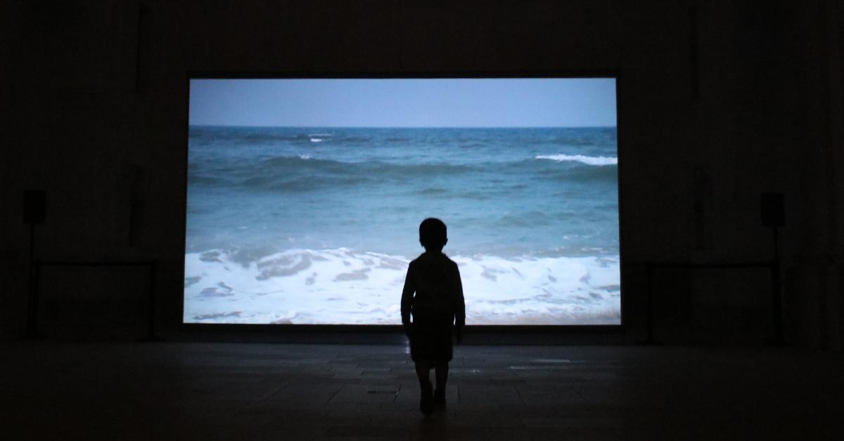 Article-waves. Küntzel. Une silhouette d'enfant se découpe dans la pénombre. Elle semble dos au photographe. Devant la silhouette, une projection. On y voit la mer.
